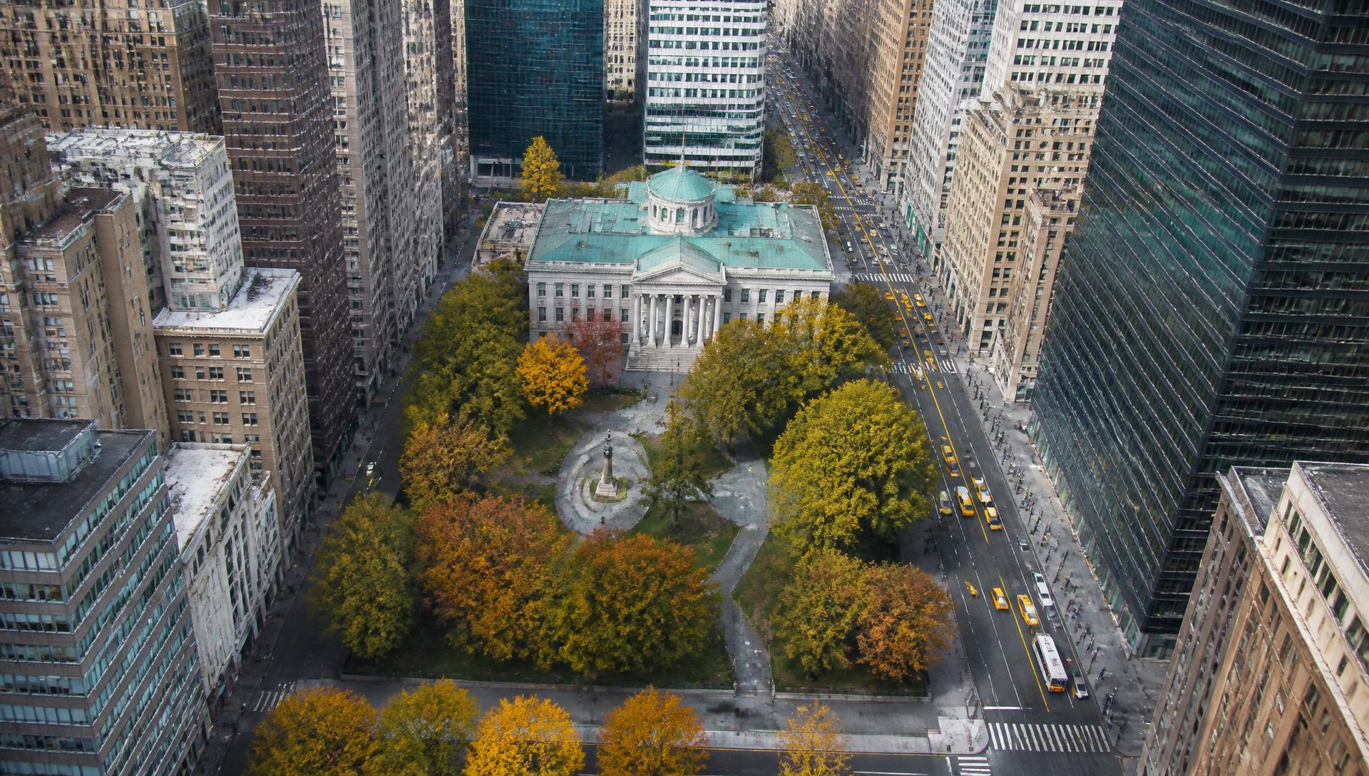 Aerial view over a leafy city park and neoclassical civic hall framed by office towers