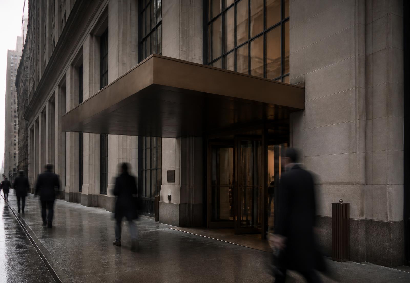 Stone building entrance under a plain bronze canopy with pedestrians passing on a wet sidewalk